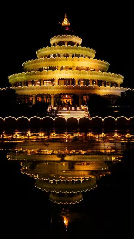 A large illuminated pavilion at night, reflected in still water.